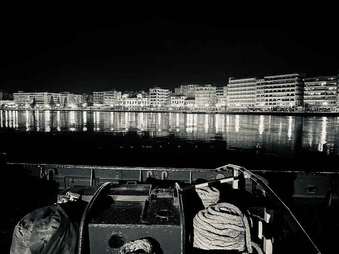 Νυχτερινή φωτογραφία της παραλιακής Χαλκίδας με φωτισμένα κτίρια που καθρεφτίζονται στη θάλασσα και σκάφος με σχοινιά στο πρώτο πλάνο